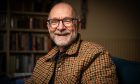 Author James Robertson at home in Angus. The picture is a head and shoulders shot of James who is wearing a checked shirt/jacket in browns and oranges. He is bald and has a greying beard and is wearing black-framed glasses and a wide, friendly smile.