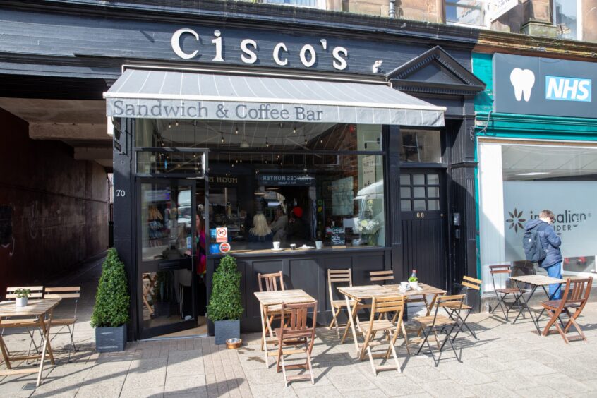 Image shows: Exterior shot of Cisco's Cafe in Stirling. The sunny shopfront has an awning and bistro chairs and tables outside.