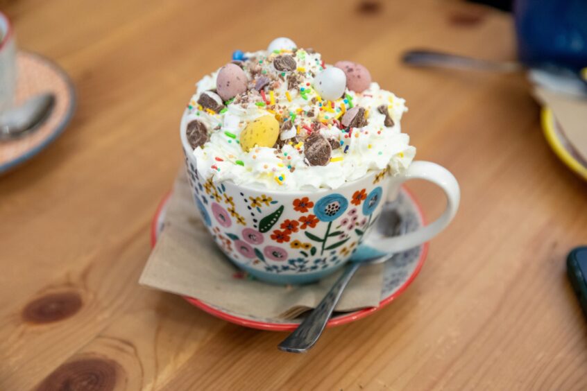 Image shows: a close up hot chocolate in a brightly painted cup with whipped cream, mini eggs and sprinkles. In The Book Nook in Stirling.