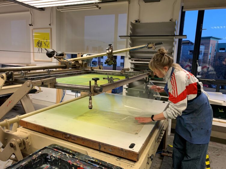 Image shows: Illustrator Kate Benzie at work in the DCA Print Studio. Kate is using a large printing press.