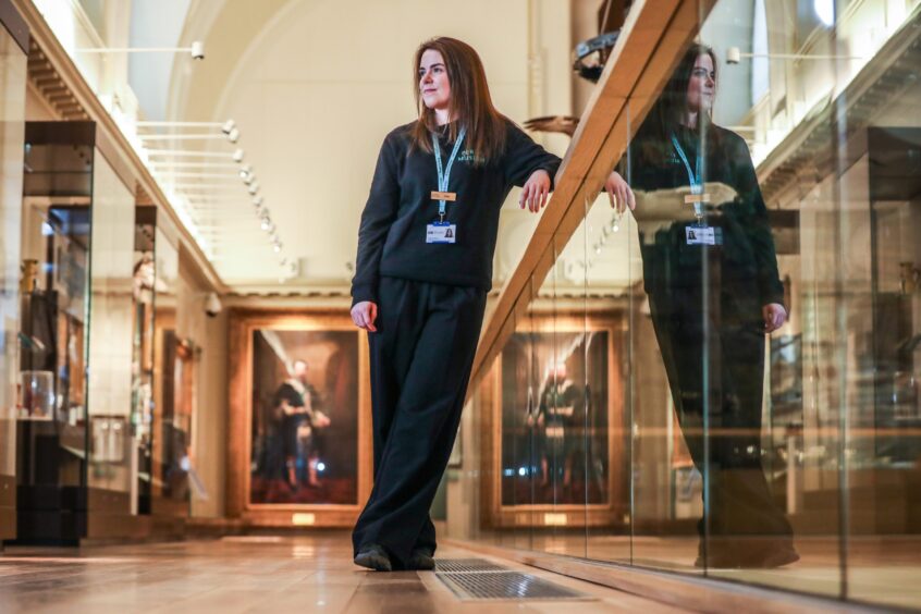 Eilidh McGlashan leaning against railing on first floor of Perth Museum surrounded by exhibits