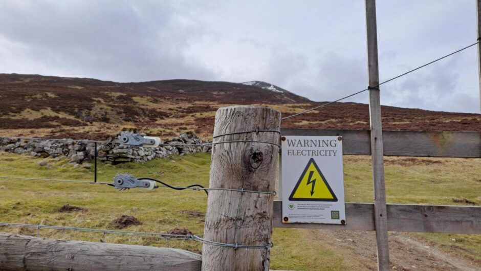 Close up of fence, with 'warning electricity' sign and Schiehallion in distance