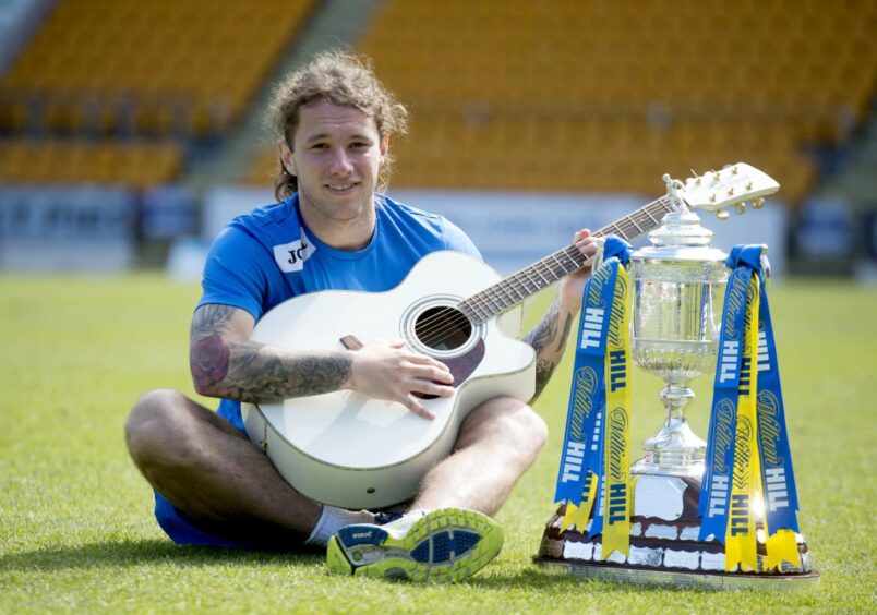 Stevie May playing a guitar on the McDiarmid Park pitch, next to the Scottish Cup trophy.