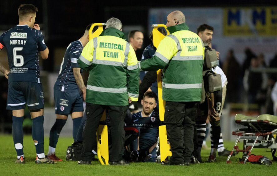 Shaun Byrne sits on the pitch surrounded by Raith Rovers team-mates and medical staff.
