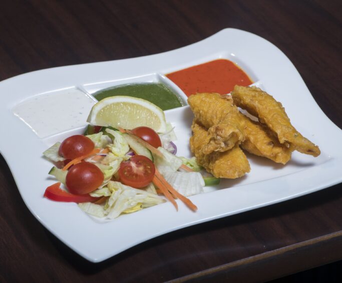 3 strips of chicken pakora on a rectangular white plate with a salad garnish of lettuce and cherry tomatoes, a slice of lemon and two dips - one green and one red.