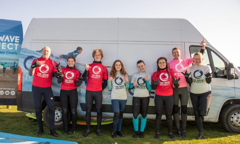 Image shows: The team of Wave Project instructors and volunteers who deliver the charity's surf club sessions.The team are wearing wetsuits and brightly coloured Wave Project t-shirts. The are standing in a row with their backs against the Wave Project branded van at West Sands, St Andrews.