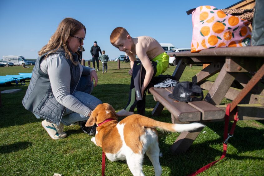 Finlay Monro gets ready for his surf club session with the help of mum Megan and a four-legged friend. Finlay is sitting on a picnic bench with Megan kneeling on the ground helping him to get into wetsuit, boots etc.