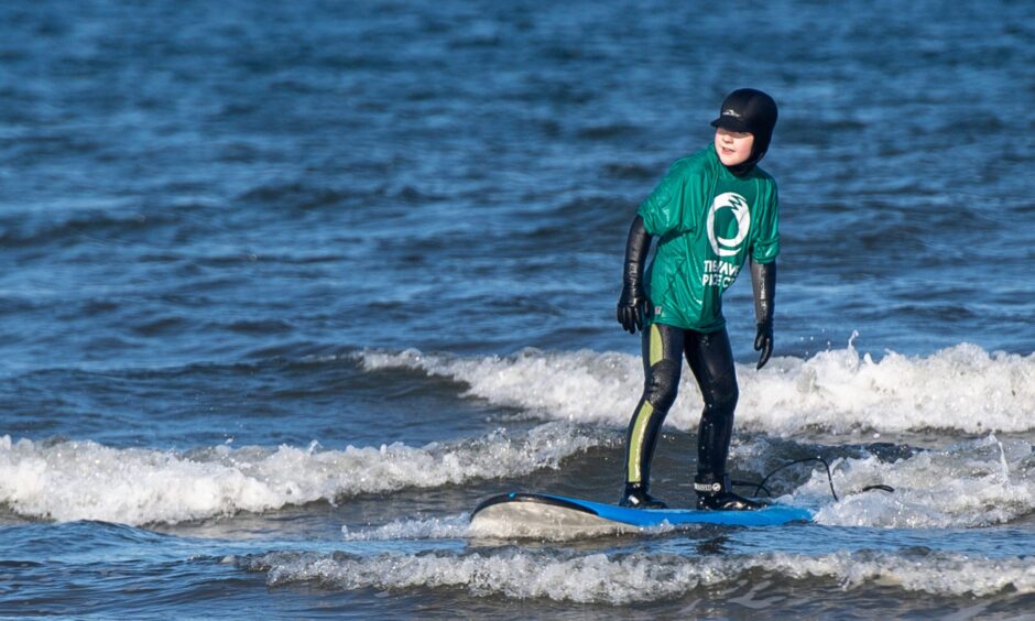 Image shows: Finlay Monro aged 10 catching a wave at West Sands Beach, St Andrews. Finlay is wearing a black wetsuit, gloves, boots and a black hood. He has a green t-shirt with the Wave Project logo on it.