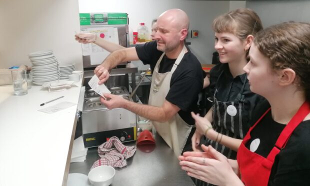 A bald man in a black T-shirt and white chef's apron looks at the orders in a commercial kitchen with two teenage girls who are training next to him.