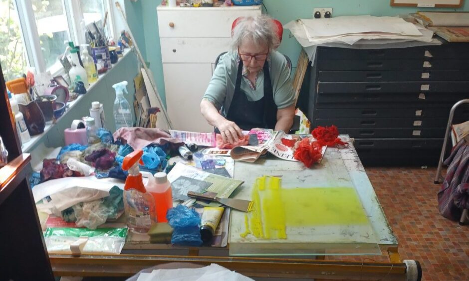 Image shows: Artist Elizabeth Shepherd in her garden studio in Crail. She is working at a desk and surrounded by colourful ink and other art materials. 