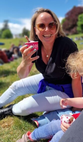 A blond woman in a black T-shirt and light blue jeans smiles at the camera while holding up a small tub of ice cream.