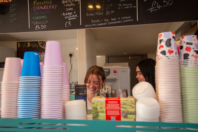 Two young women behind a counter smiling while serving customers in a café.