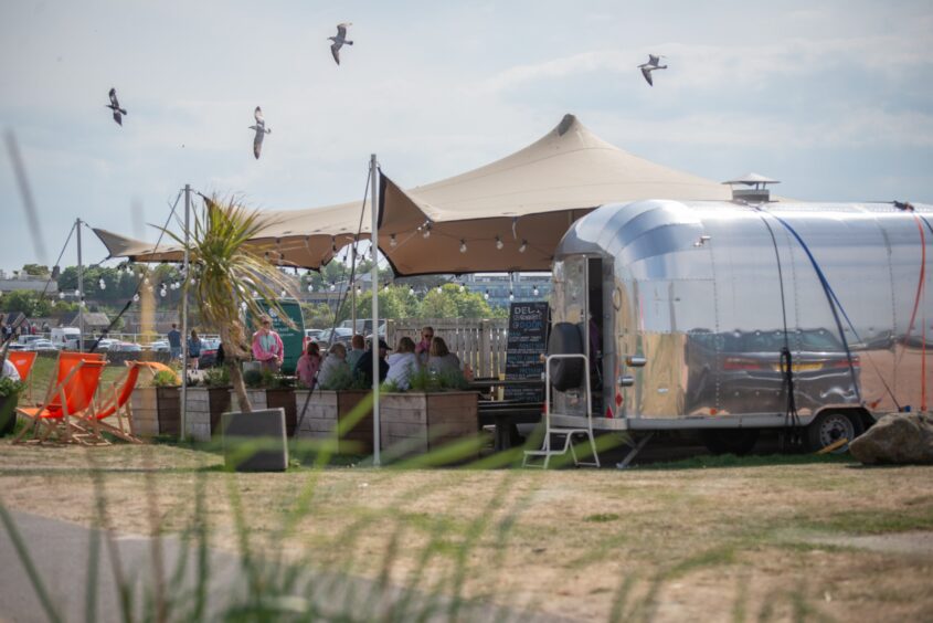 A peaked canopy outside a café with picnic tables beneath and a silver food truck by a beach with sea gulls flying overhead.