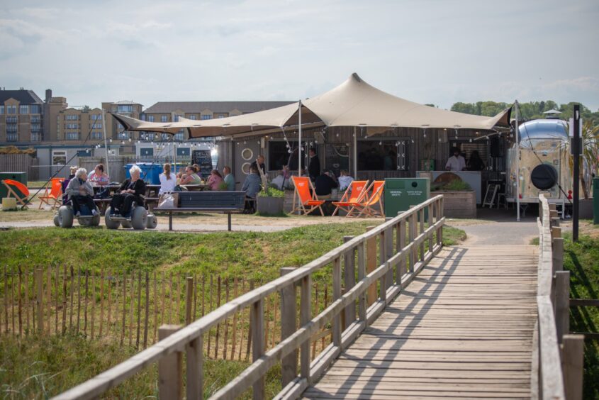 A wooden walkway leads from the beach to a café with a large canopy stretched over picnic tables.