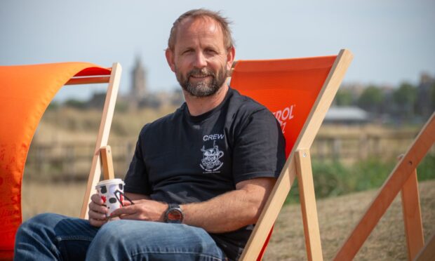 A man in a black t short and blue jeans sits on a bright orange deck chair with the St Andrews skyline behind him