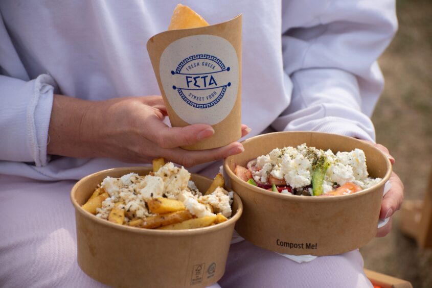 A woman in a white sweatshirt holds three cardboard dishes on feta-covered fries and a Greek salad.