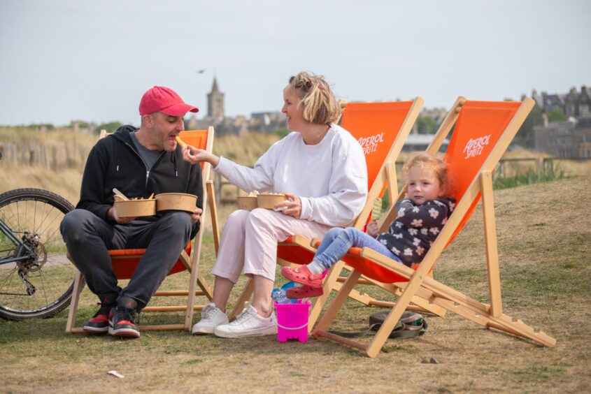 A blond woman sitting on an orange deck chair feed a piece of food to her husband in a red cap and black hoodie while their young daughter sits on another orange deckchair next to them.