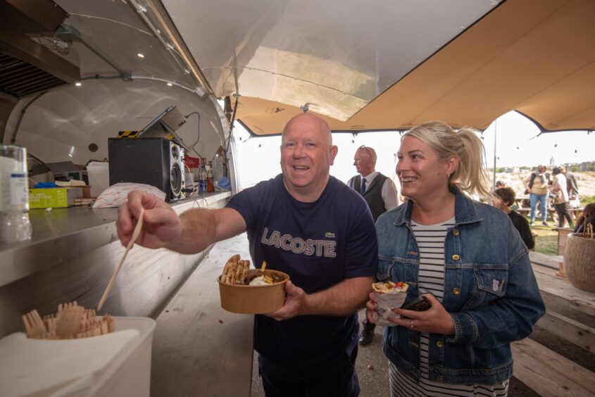 A bald man smiles at the camera while reaching for cutlery to go with his food outside a silver foodtruck.