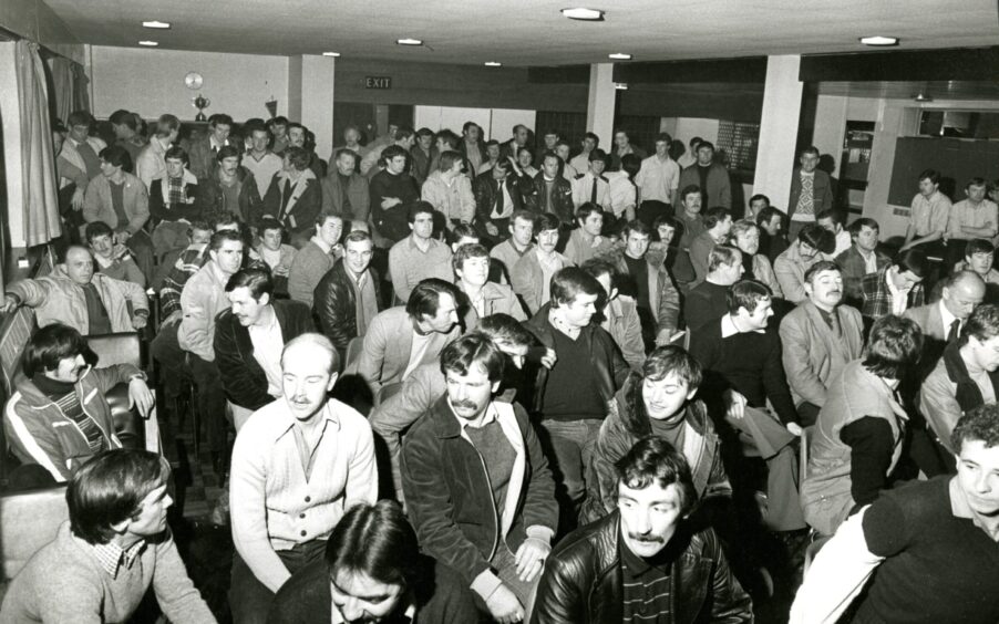 Men standing a the back of a hall and more seated in front of them, as Tayside branch of firefighters are pictured before their meeting. 