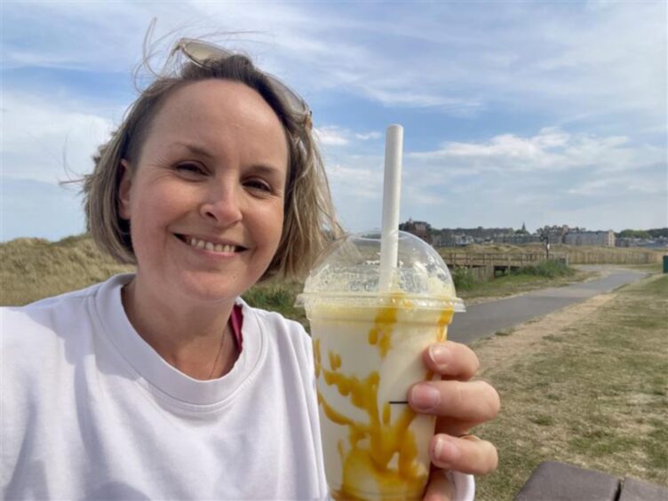 A smiling blond woman in a white sweatshirt hold a plastic milkshake cup with a straw - the milkshake is yellow and white with cream on top.