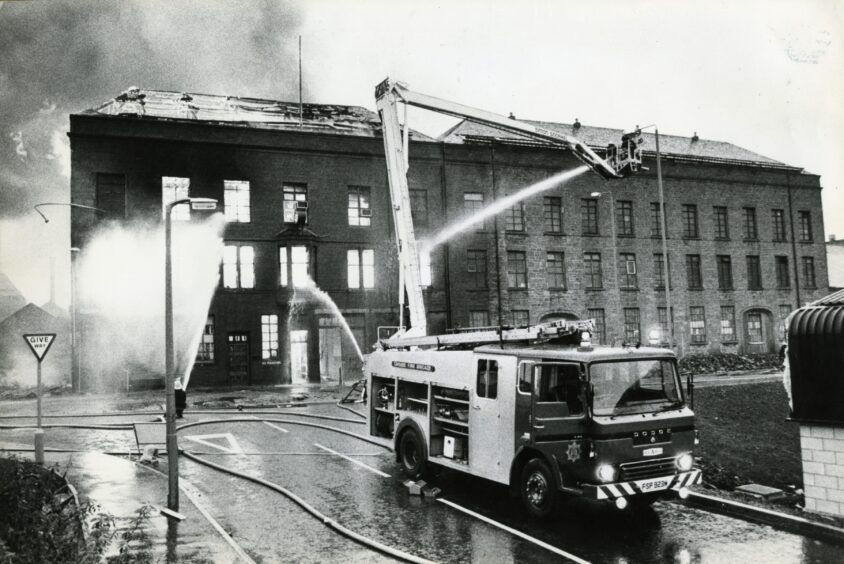 a fire engine is in the foreground, which is elevating firefighters so they can hose down the burning building 