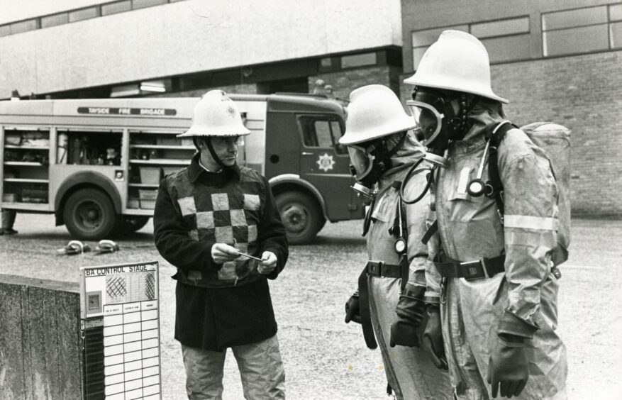 Three Tayside Fire Brigade firefighters during training, two of whom are wearing breathing apparatus. A fire engine is parked in the background 