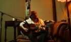 Image shows: Singer-songwriter Blair Davie. Blair is sitting on a sofa playing guitar there is a microphone in the foreground.