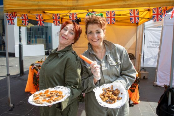 Two women in khaki holding plates of food in front of stall at Perth VE day event