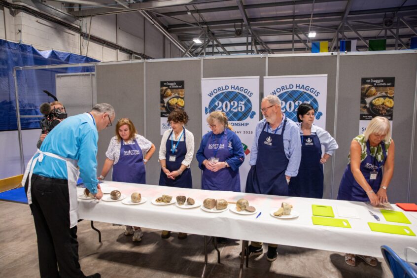 Row of people in blue aprons standing behind table with several haggises on plates in front of them