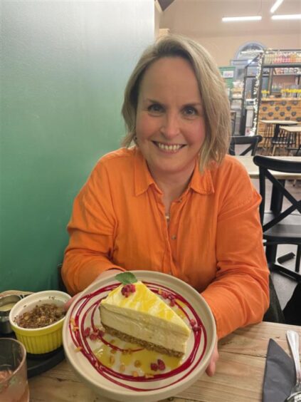 A blonde lady in an orange shirt holds a plate containing a large slice of baked cheesecake while smiling at the camera.