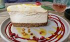 A large slice of creamy baked cheesecake on a white plate with circles of red syrup on the plate and topped with a layer of creamy mousse and a red berry.