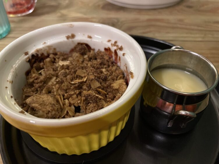 A black plate with a yellow ramekin of fruit crumble next to a silver jug of custard.