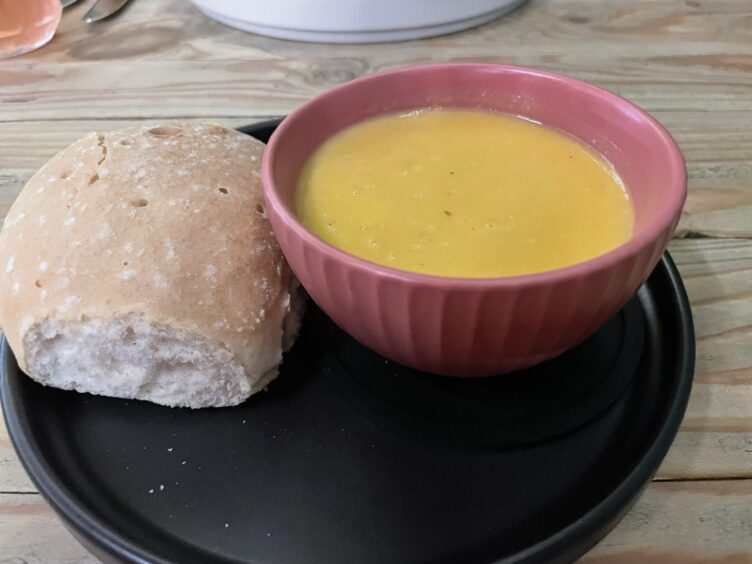 A bowl of yellowy butternut soup in a small pink bowl with a roll next to it.