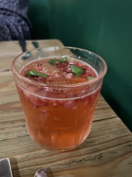A small glass of pink fizzy cocktail with bits of raspberry and mints floating in it on a wooden table.