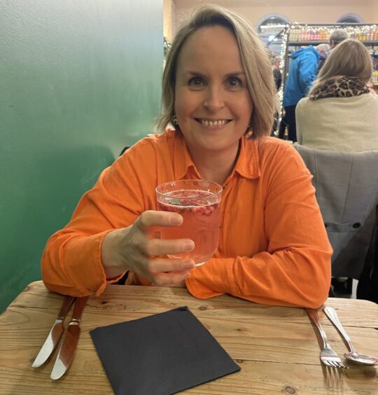 A blond women in an orange shirt smiles at the camera while holding a small glass containing a pink rose fizz cocktail.