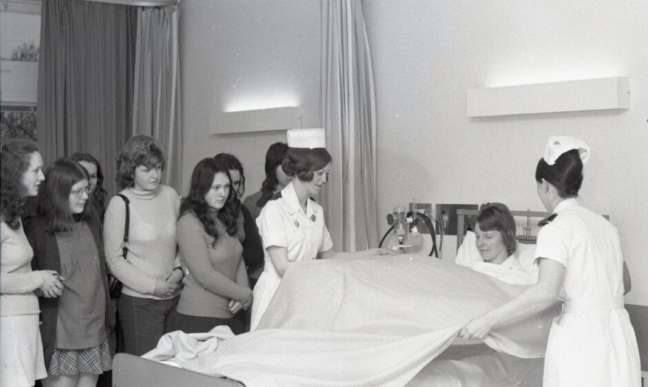 a group of women gather round to watch as two nurses attend to a female patient in bed