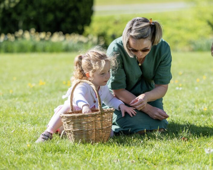 A blond woman in a green dress crouches on the grass with a young girl in a pink dress and they examine the grass.