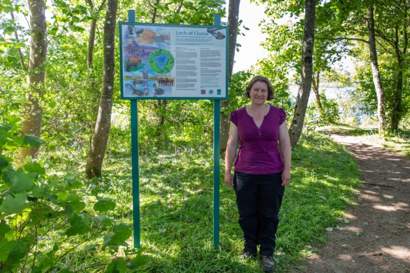 Janice Willsher standing next to information board in trees beside loch of Clunie