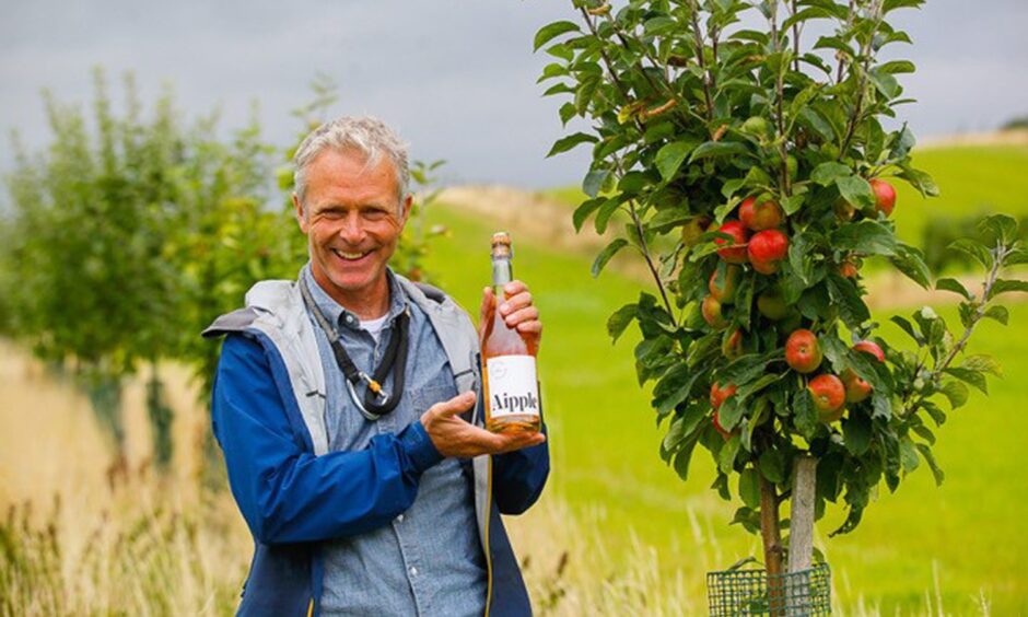 A man holding a bottle of cider and standing by an apple tree.
