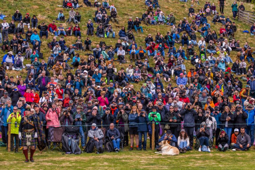 Large crowd of people seated on grassy slope watching highland games