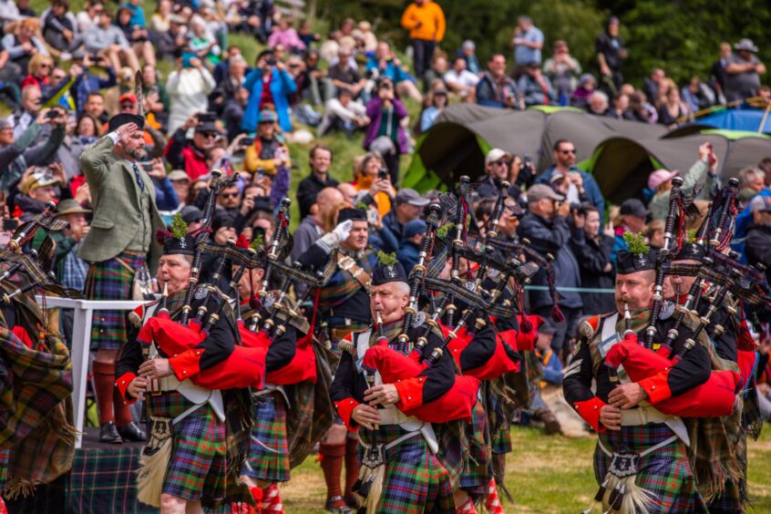 Pipe band playing bagpipes as they march past man in Highland dress saluting