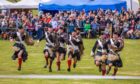 A line of men in full Highland regalia run towards the camera in a grass areana.