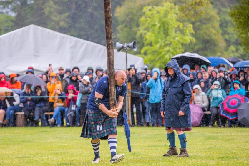 Heavyweight athlete in kilt preparing to toss caber watched by crowd in heavy rain
