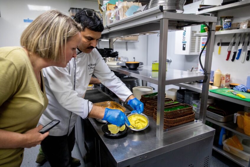 A blond woman in a green top watches an Indian chef making a coconut sauce.