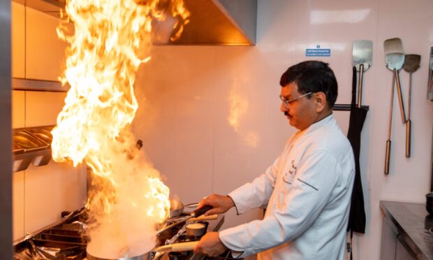 An Indian chef in chef's whites shakes a pan calmly while flames fly up in to the air.