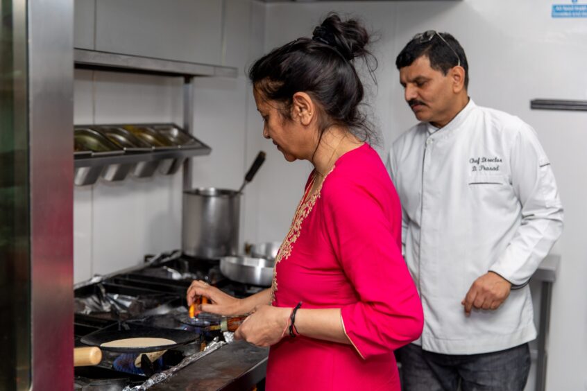 An Indian woman in a pink dress makes chapati over a flame on a commercial hob while the chef stands next to her.