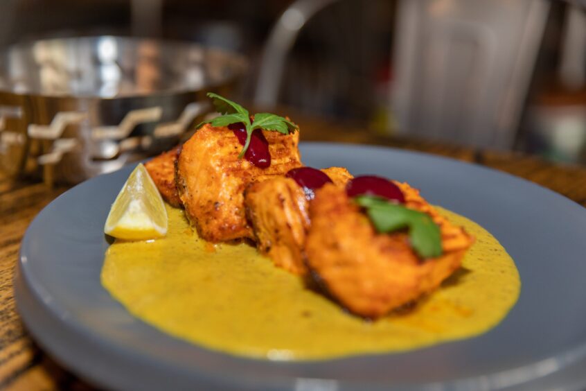 Pieces of cooked marinated salmon on a coconut sauce with blueberry sauce and fresh coriander on top of the fish and a wedge of lemon the side.