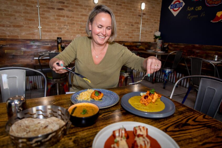 A blond woman in a green top smiles as she tucks into various Indian dishes.
