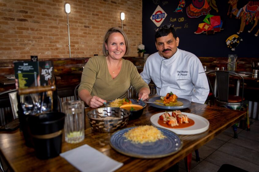 A blond woman in a green top sits at a table of Indian food next to an Indian man in a white chef's jacket.