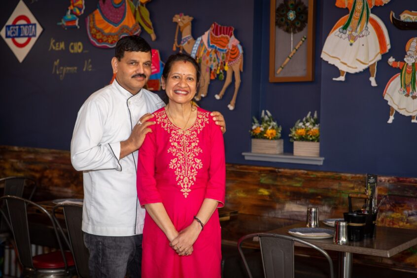 An Indian man in a white chef's jackets stands smiling next to his wife who wears a long bright pink dress in their Indian restaurant.